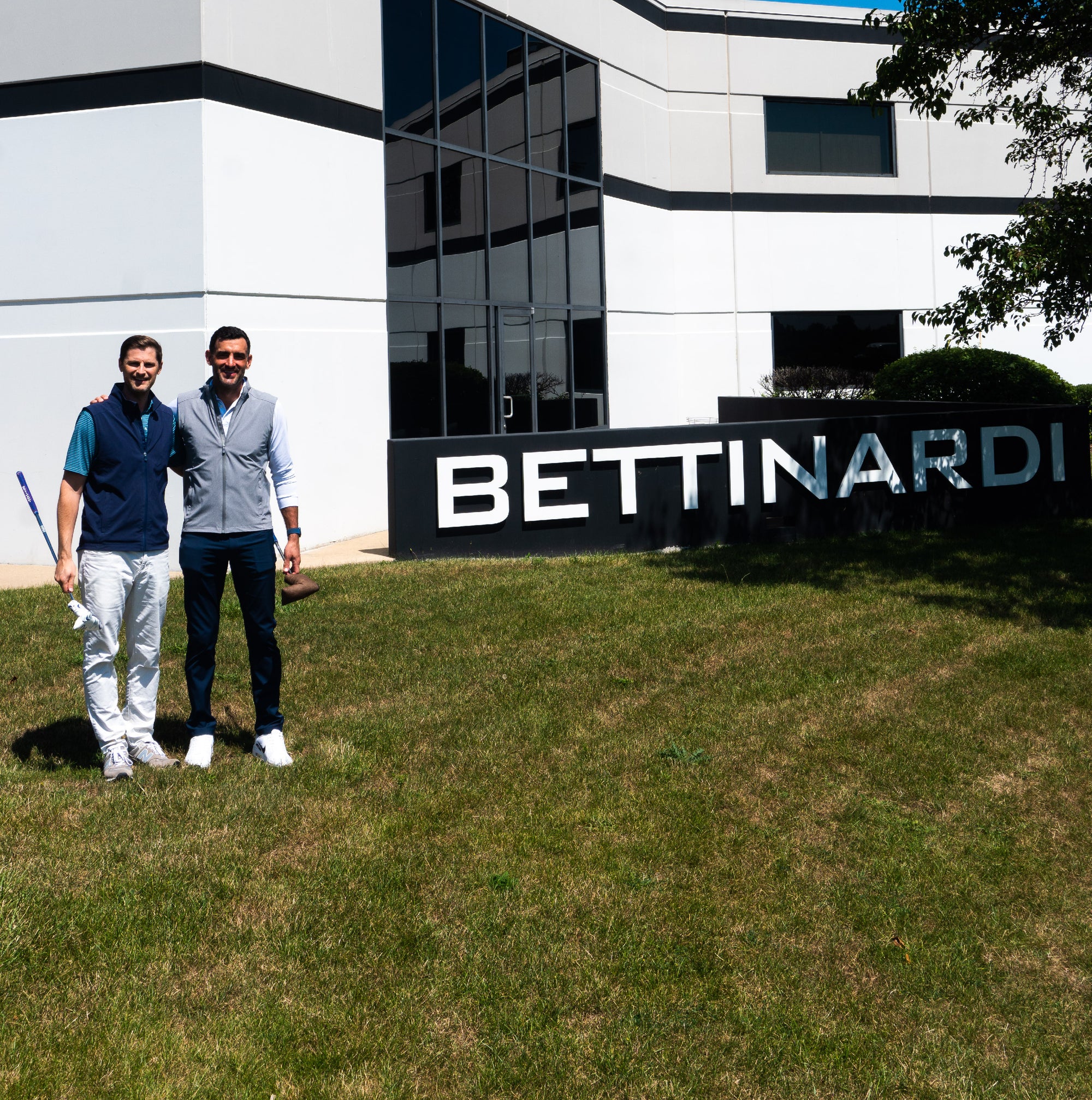 Two men, one in a navy vest and one in a gray vest, smiling on the lawn in front of the "BETTINARDI" building sign.