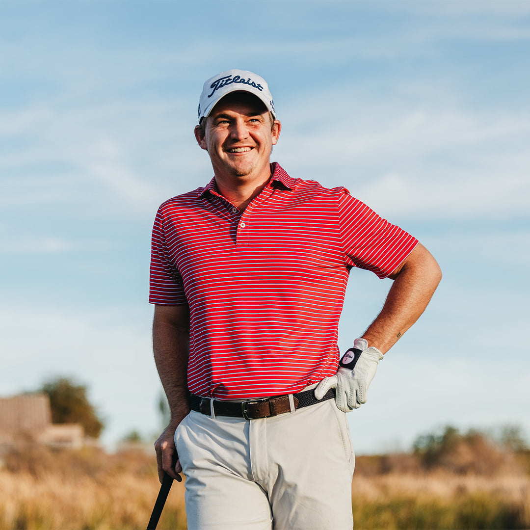 Smiling golfer in a white Titleist cap, red striped polo and stone pants with a brown belt, hand on hip and a club in the other hand.