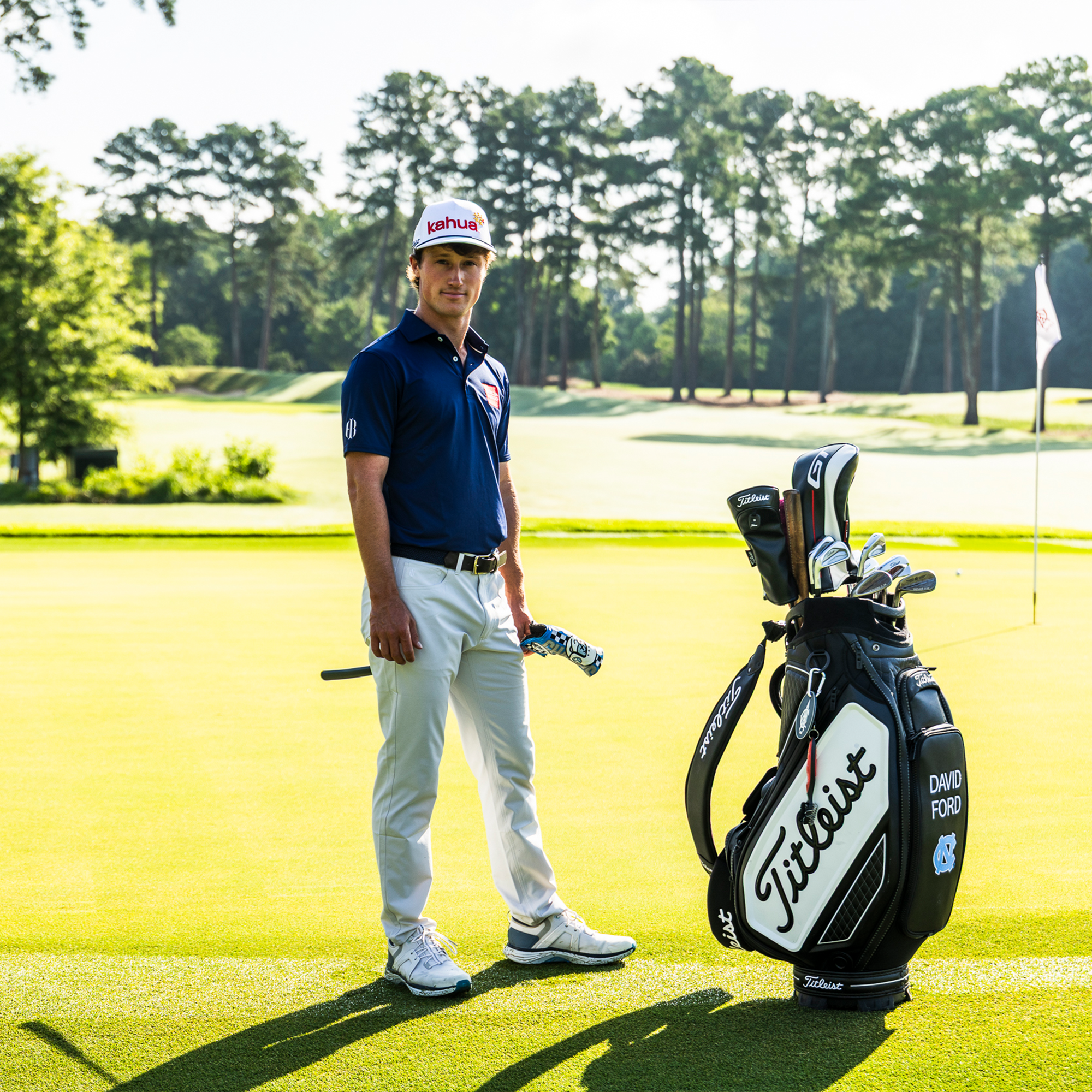Golfer in a navy polo, white pants and logo cap standing by a Titleist tour bag marked "DAVID FORD" on a green.