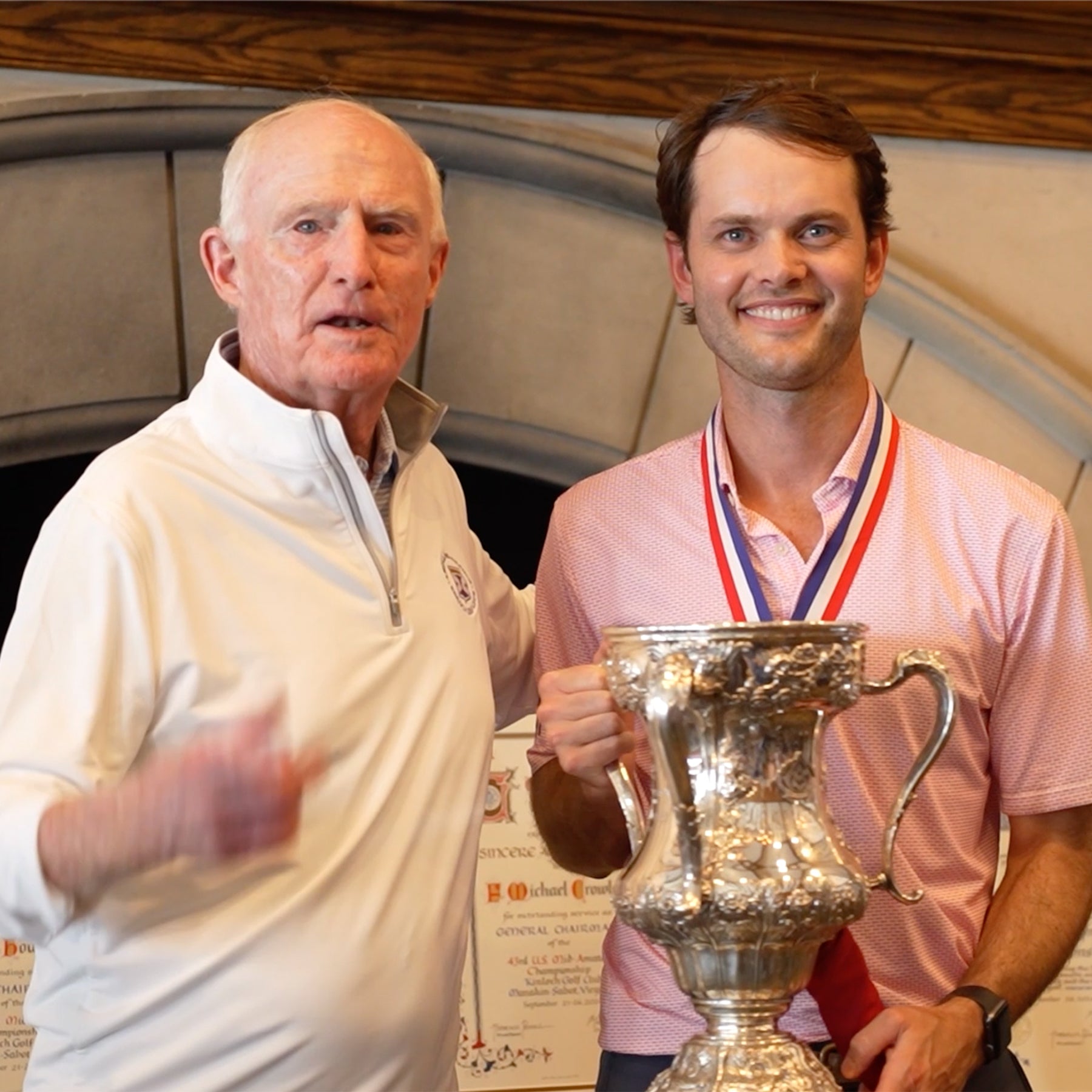 Smiling golfer in a pink polo wearing a medal and holding a silver cup, beside an older man in a white quarter-zip.
