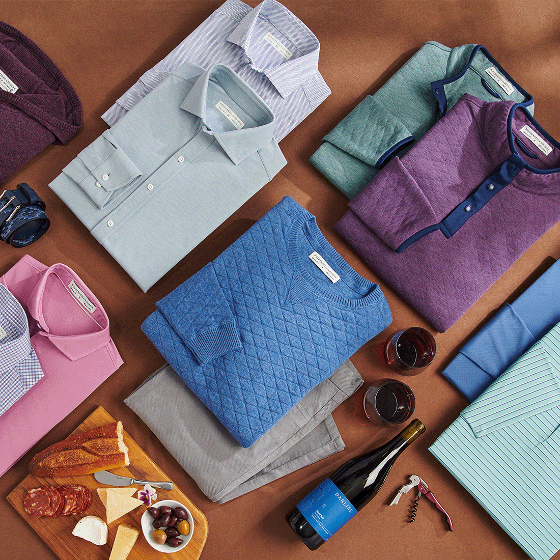 Flat lay of folded button-downs, quilted pullovers and a henley in blue, purple and green with chinos, wine and a cheese board.