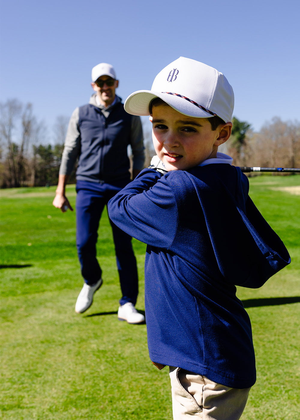 Young boy in a navy pullover and white cap taking a golf swing, with a man in a navy vest smiling behind him.