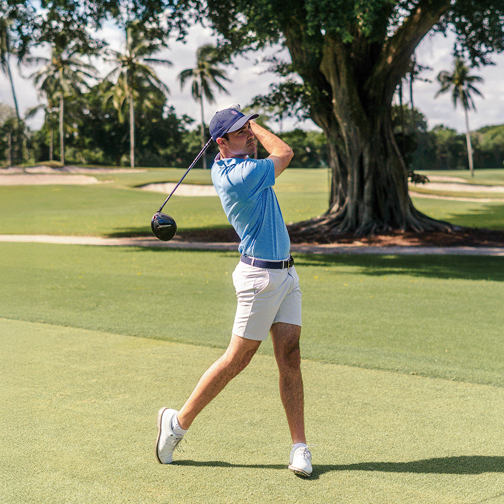 Golfer in a blue polo, white shorts and navy cap finishing a drive on a tropical course with palm trees and a large banyan tree.