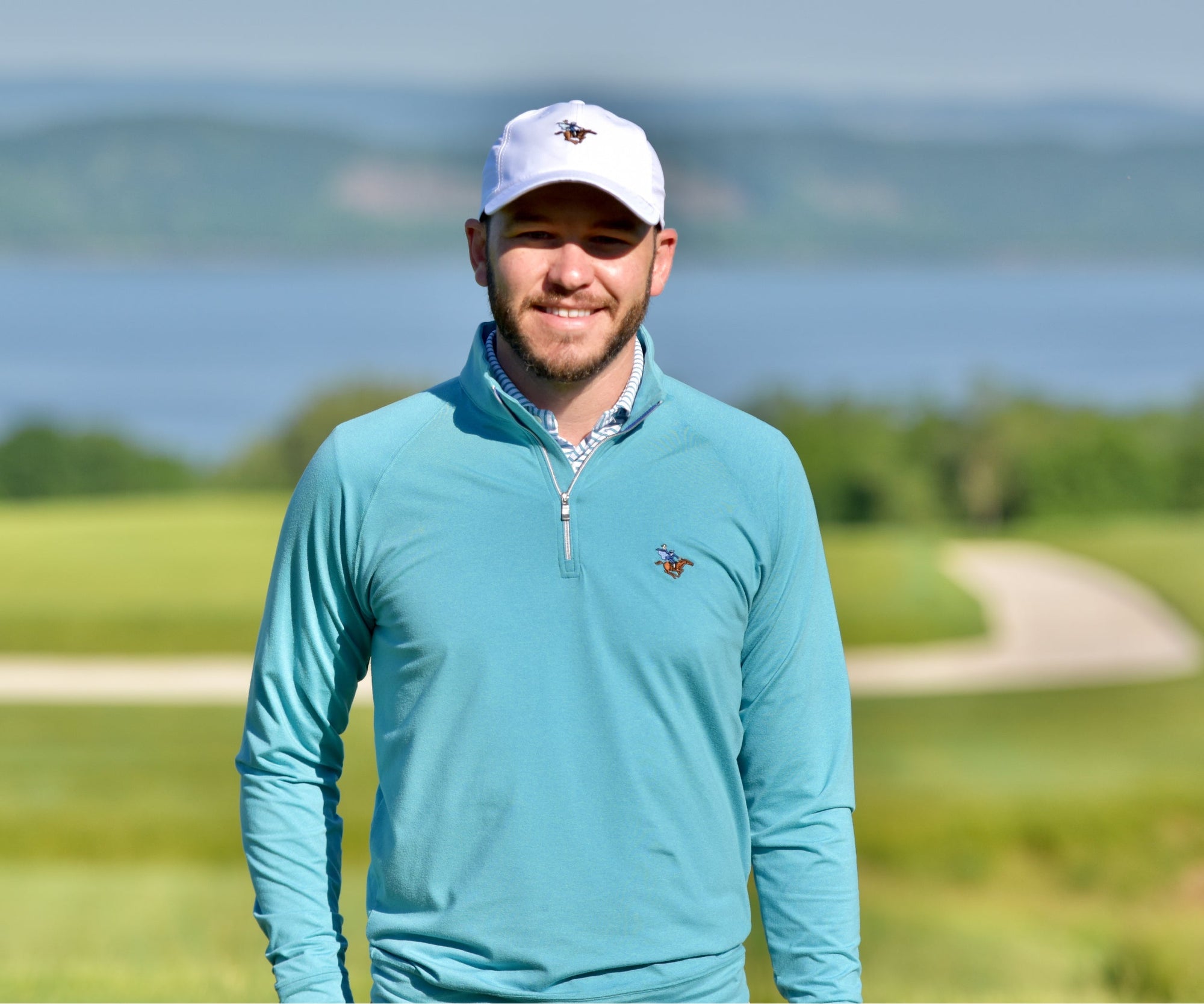 Smiling bearded man in a white cap and teal quarter-zip standing on a golf course overlooking a lake.