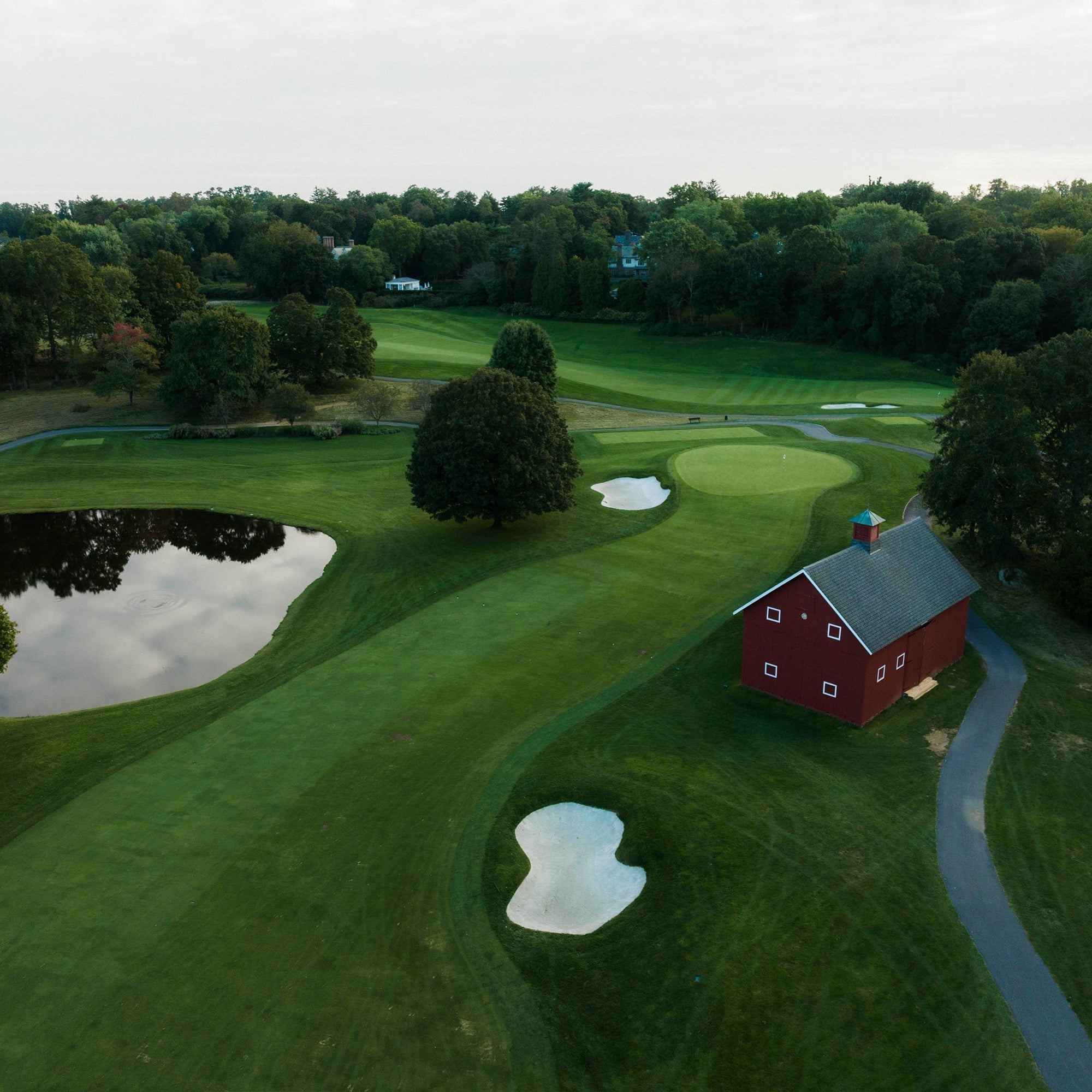 Aerial view of a golf hole with a green, bunkers, a pond and a red barn surrounded by trees.