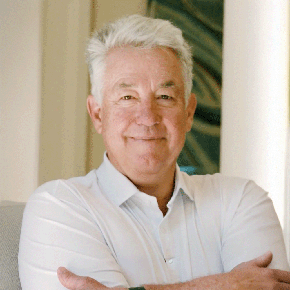 Smiling older man with gray hair in a white shirt with arms crossed, seated indoors.