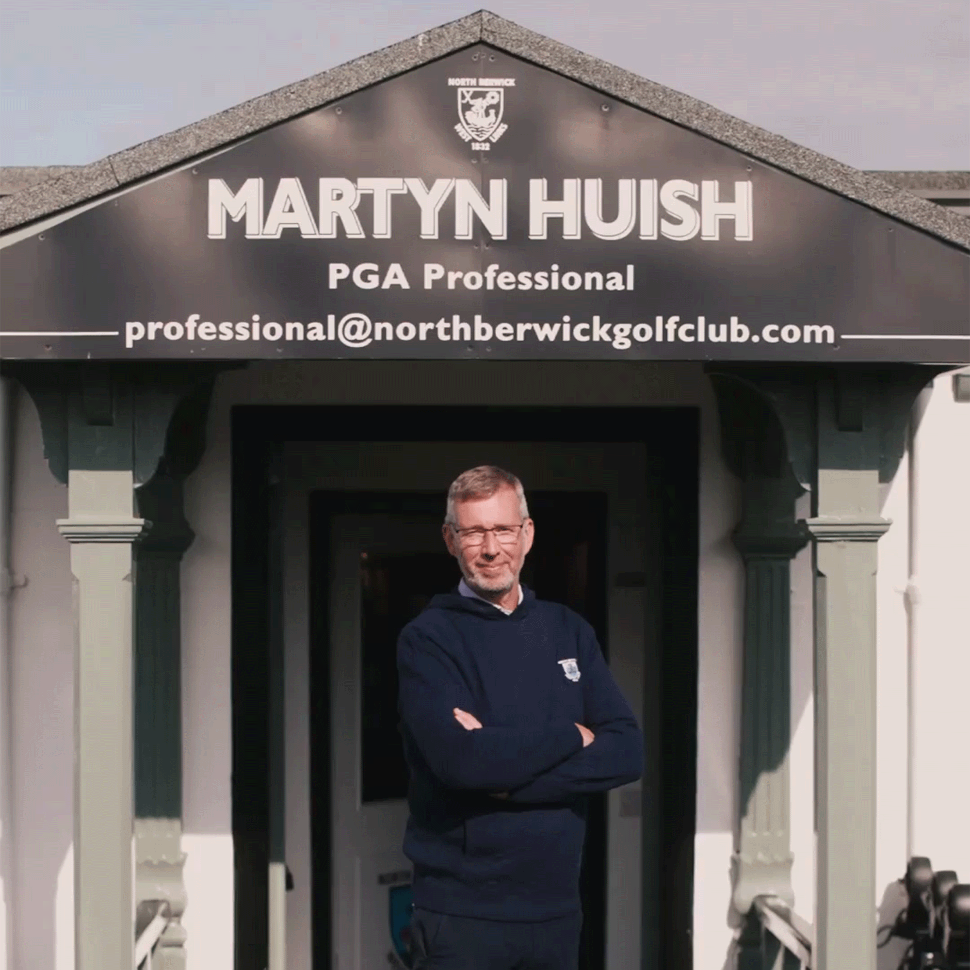Smiling man in glasses and a navy sweater with arms crossed under a sign reading "MARTYN HUISH" and "PGA Professional".