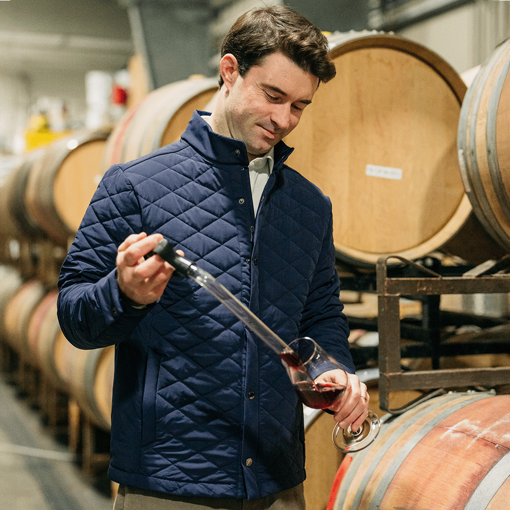 Smiling man in a navy quilted jacket pouring red wine from a barrel sample into a glass in a cellar with oak barrels.