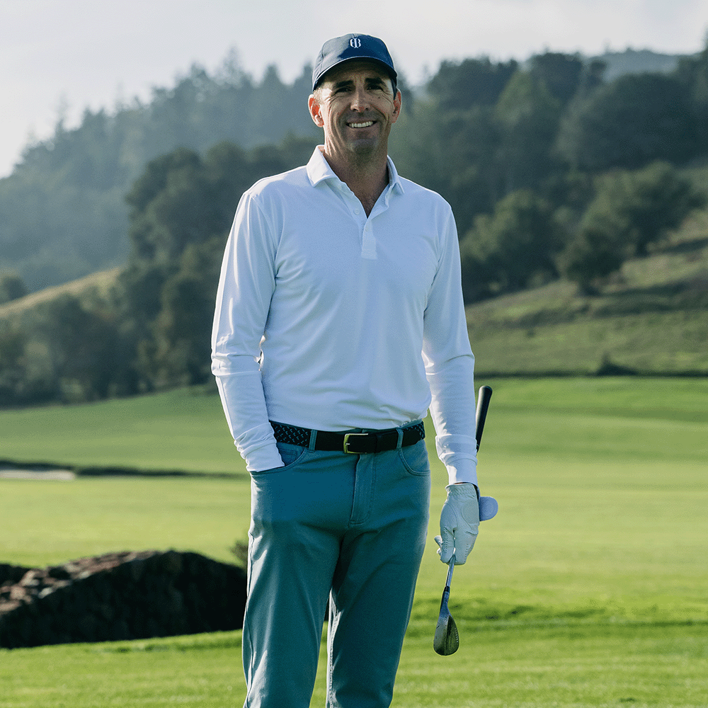 Smiling golfer in a navy cap, white long-sleeve polo, woven belt and teal pants holding a wedge on a fairway with wooded hills behind.