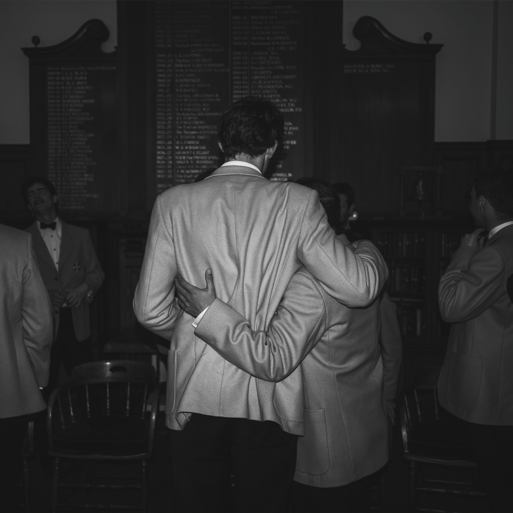 Black and white photo of men in blazers embracing in a wood-paneled hall, with honor boards on the wall behind them.