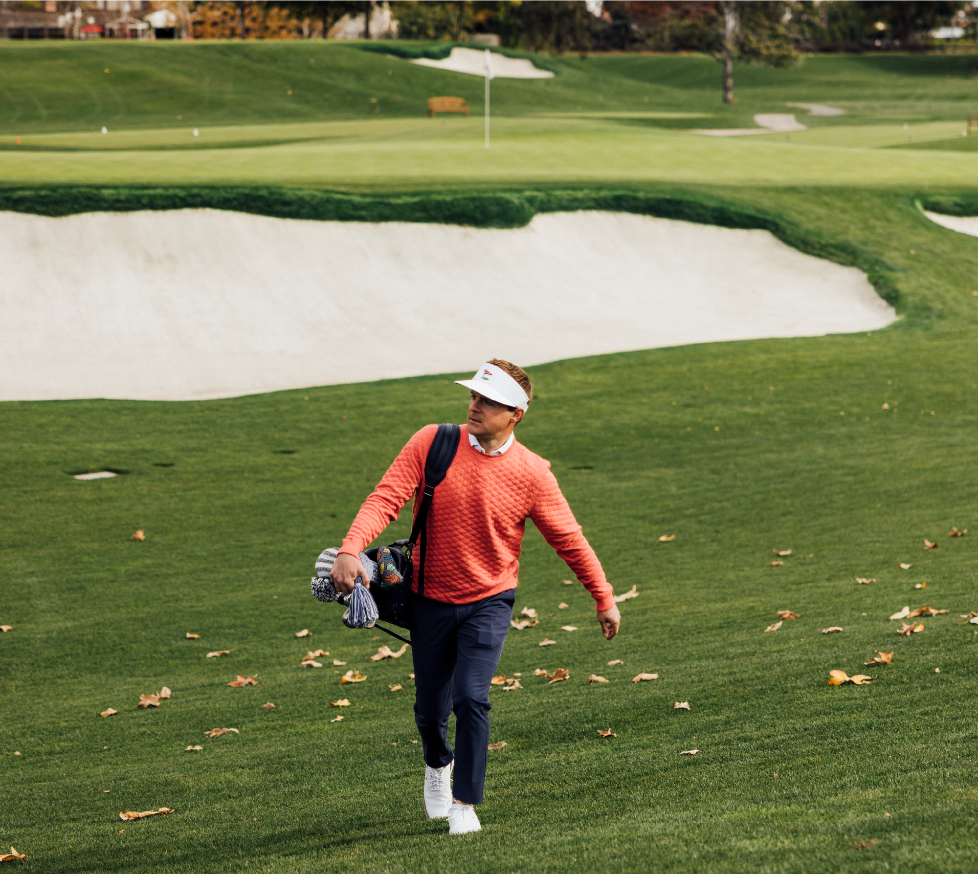 Golfer in a white visor, coral quilted sweater and navy pants carrying a bag across a green with fallen leaves and a bunker behind.