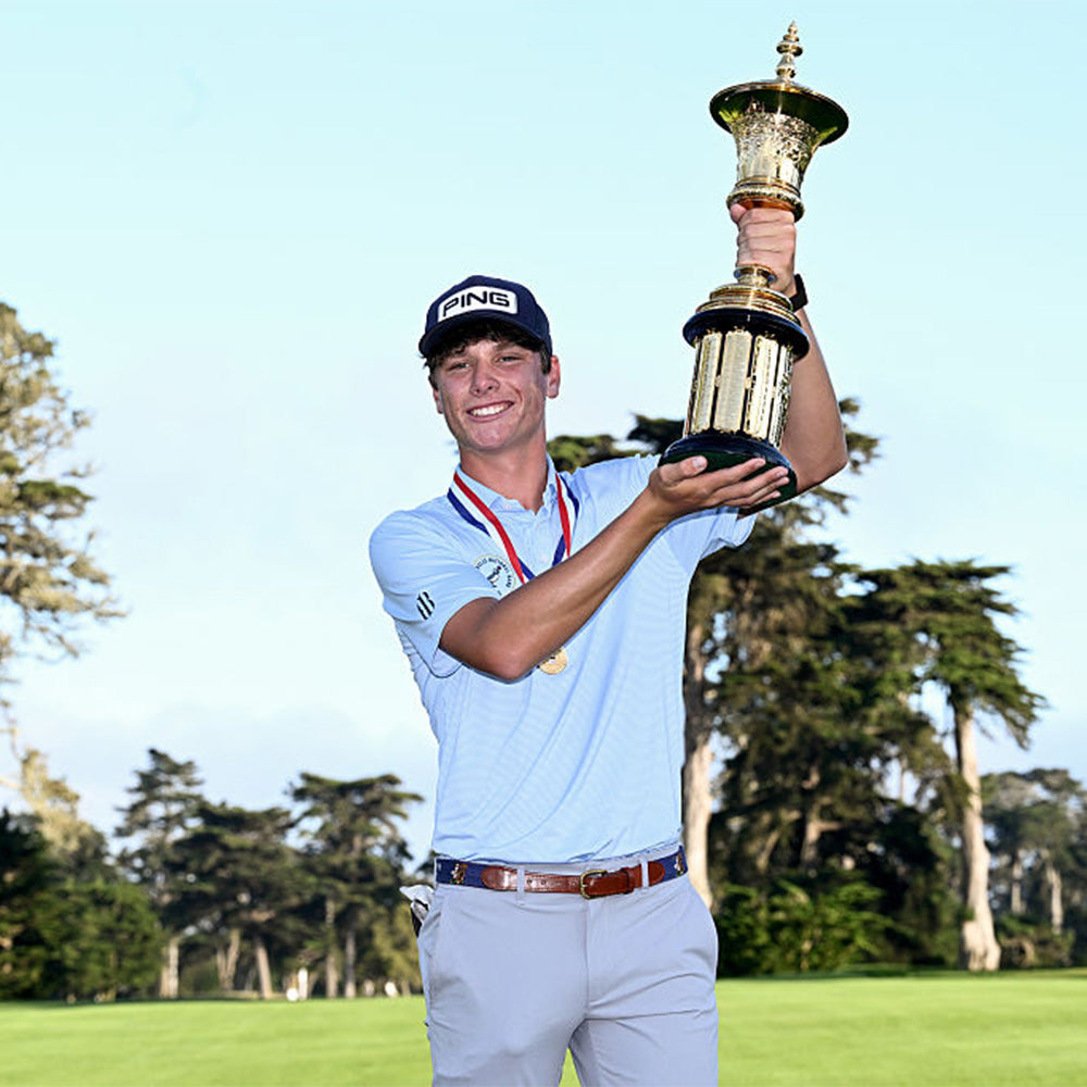 Smiling young golfer in a PING cap and light blue polo wearing a medal and raising a gold trophy on a course.
