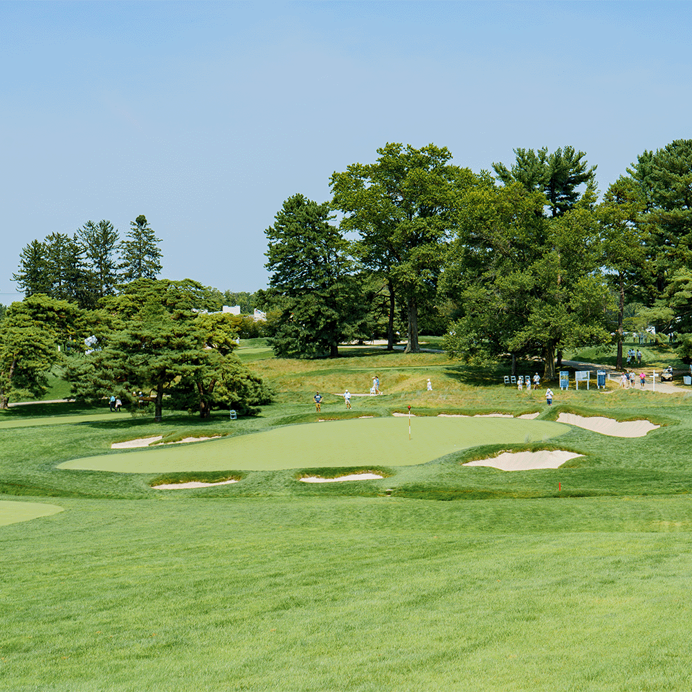 Green golf course hole with a putting green, sand bunkers and tall trees, with spectators walking in the distance.