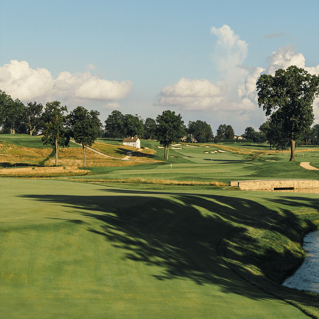Rolling golf course hole with a green in the distance, a white outbuilding, a stone bridge and shadows across the fairway.