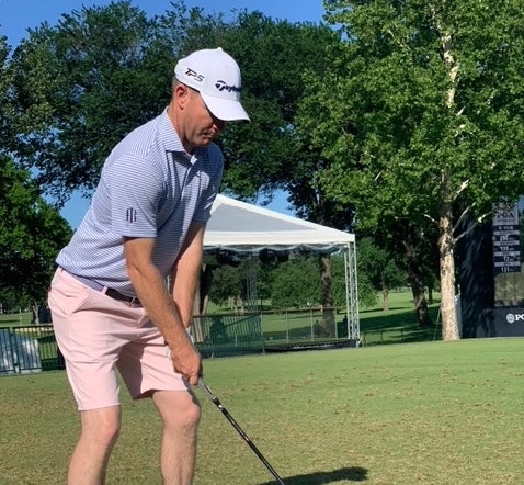 Golfer in a white cap, blue striped polo and pink shorts addressing the ball on a range with a white tent behind him.