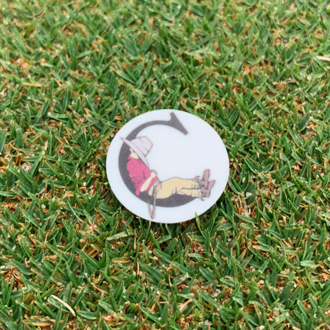 Round white ball marker showing a cowboy lounging inside a large letter C, resting on short grass.