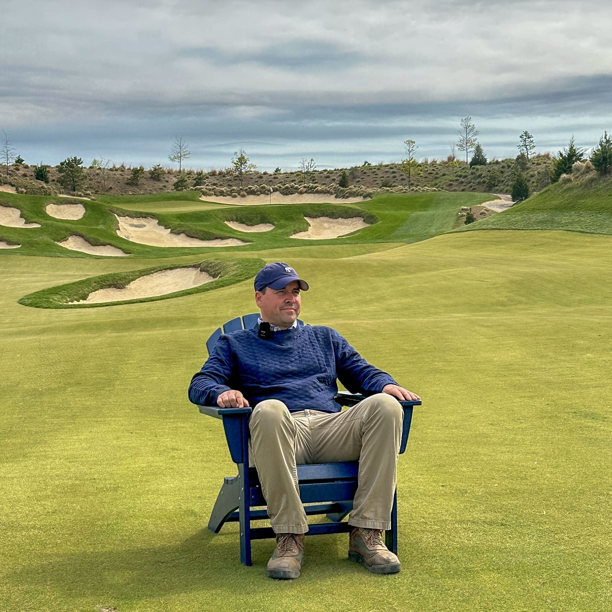 Man in a navy cap and sweater and khakis seated in a blue chair on a links fairway with sand bunkers behind him.