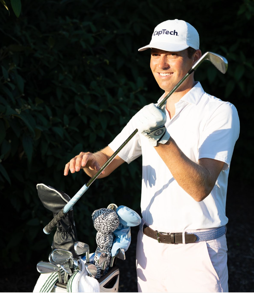 Smiling golfer in a white "CapTech" cap and white polo holding an iron beside a golf bag with colorful headcovers.