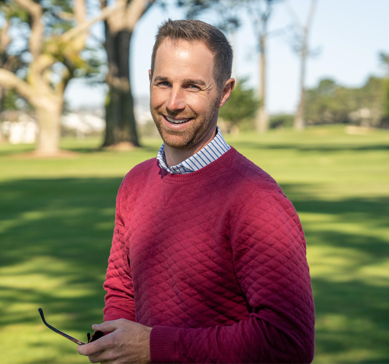 Smiling man in a red quilted crewneck sweater over a striped collar, holding sunglasses on a sunny golf course.