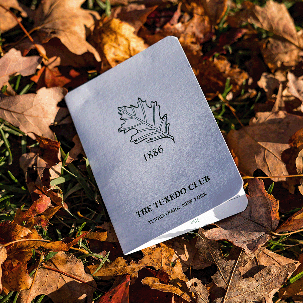 Small gray booklet reading "THE TUXEDO CLUB", "TUXEDO PARK, NEW YORK" and "1886" with a leaf crest, lying in autumn leaves.