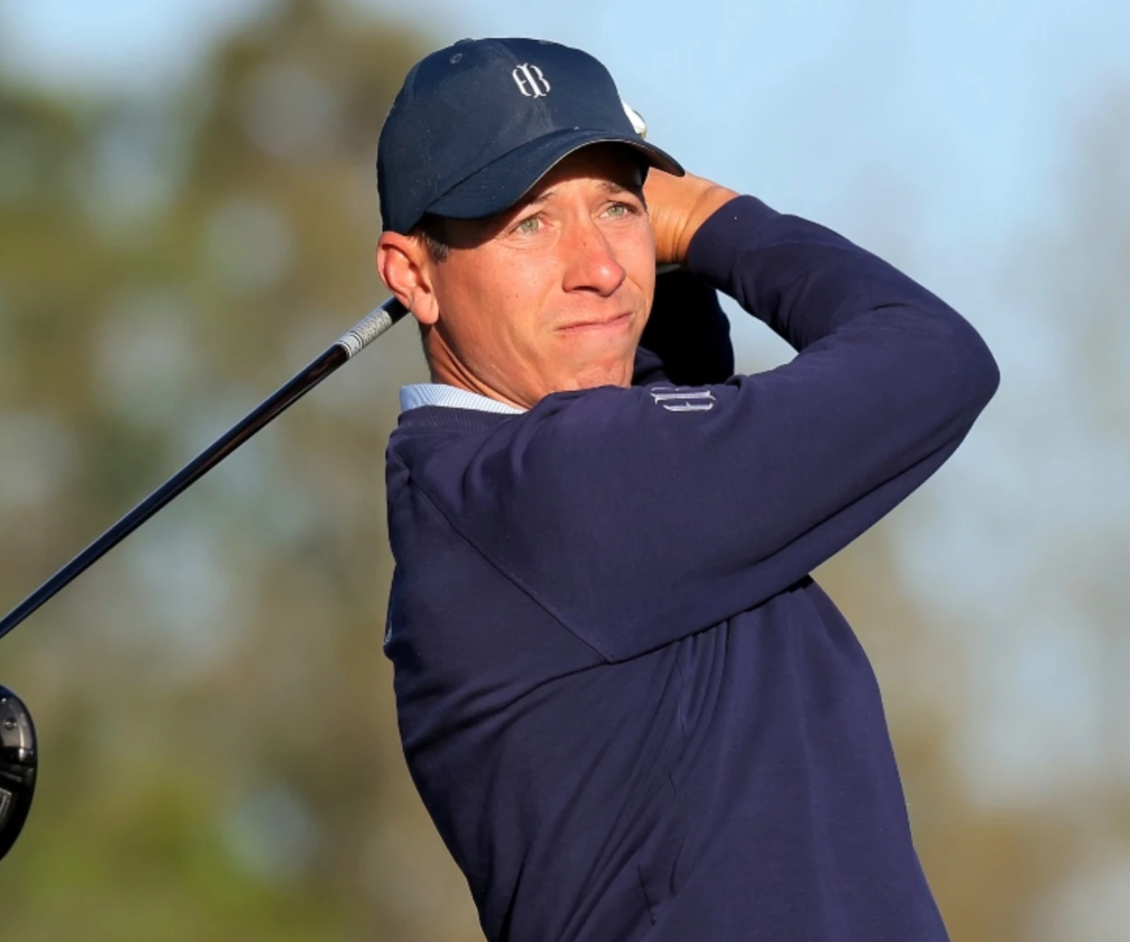 Golfer in a navy cap and navy sweater with a small logo finishing a tee shot, club behind his head.