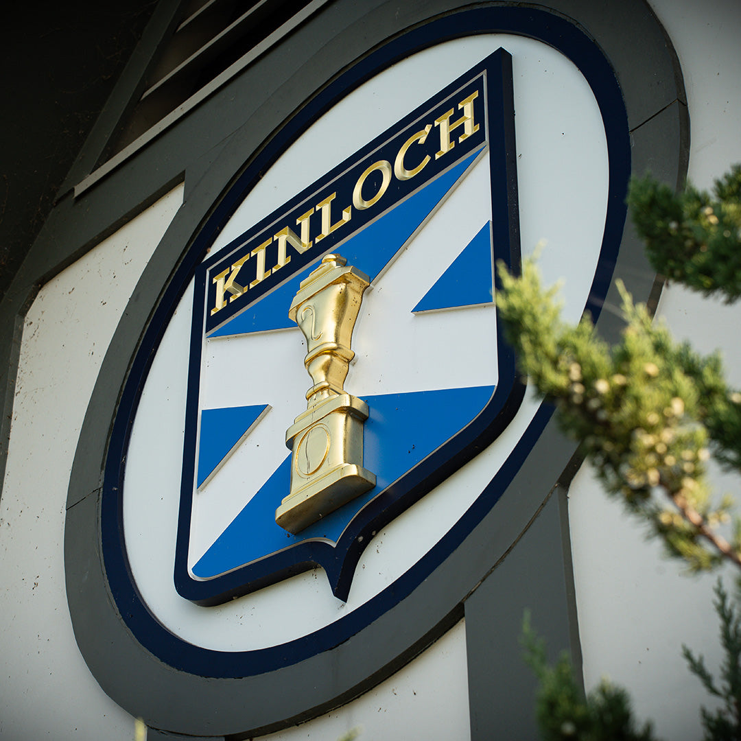 Round sign with a blue and white shield showing a gold urn below the gold word "KINLOCH", beside a cedar branch.