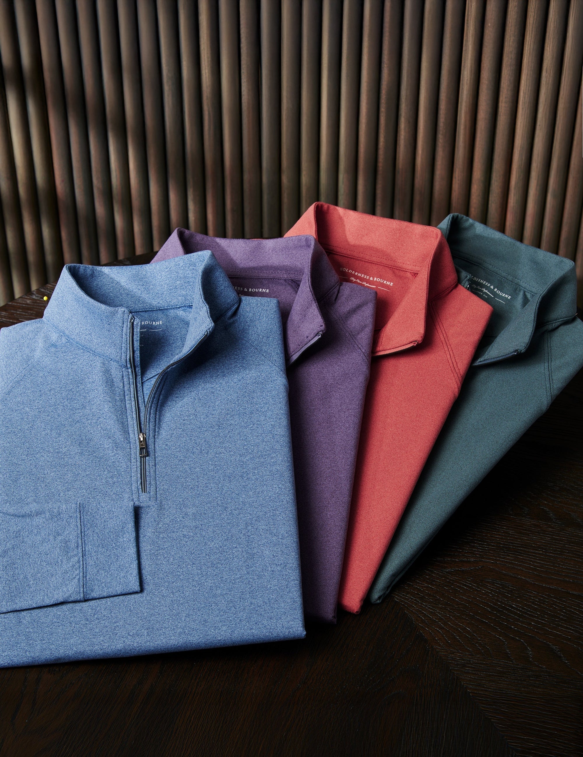 Four folded quarter-zip pullovers in blue, purple, coral and dark green fanned out on a dark wood table against a slatted wall.