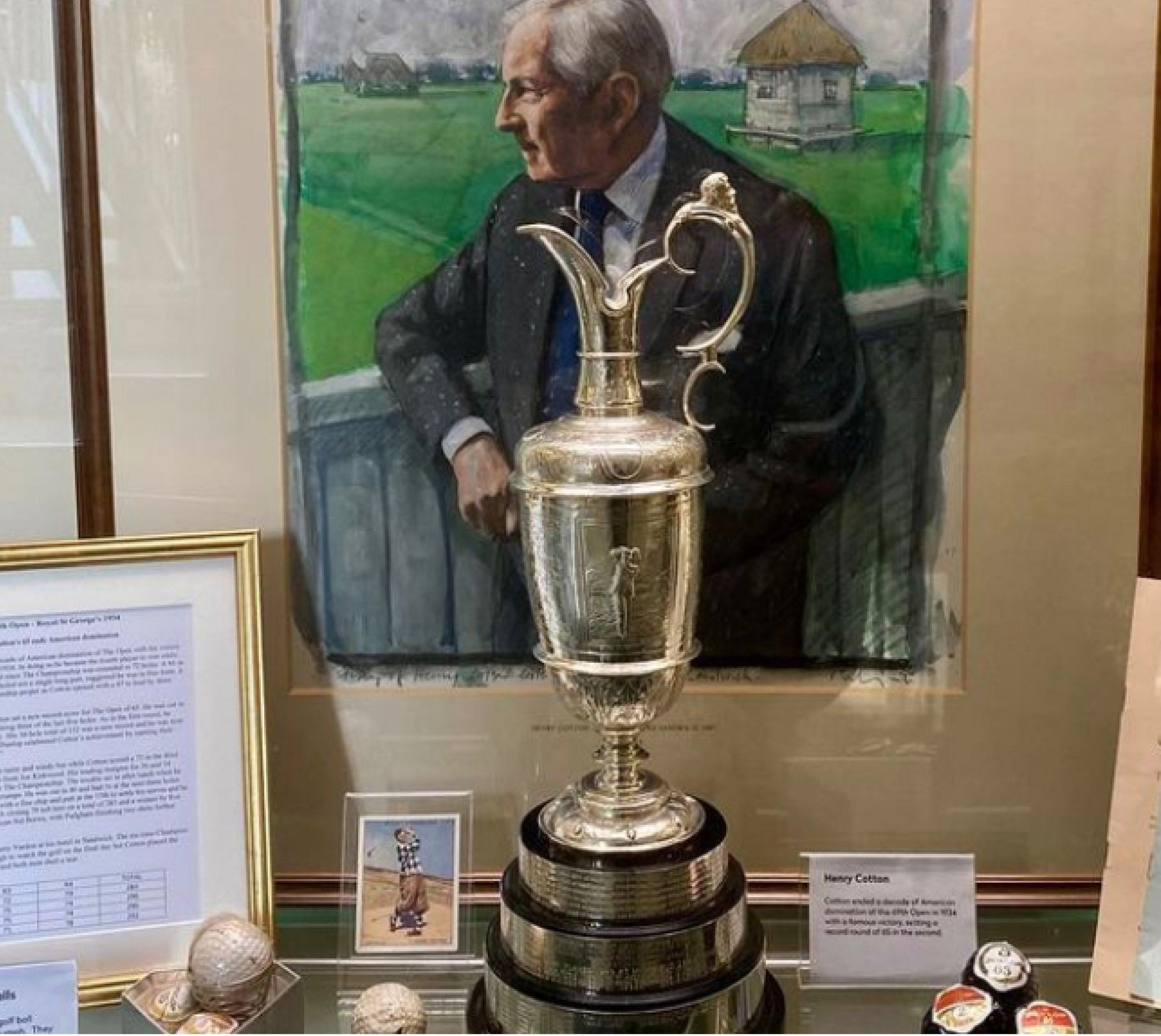 Silver claret jug trophy on display in front of a framed portrait, beside a placard that reads "Henry Cotton".