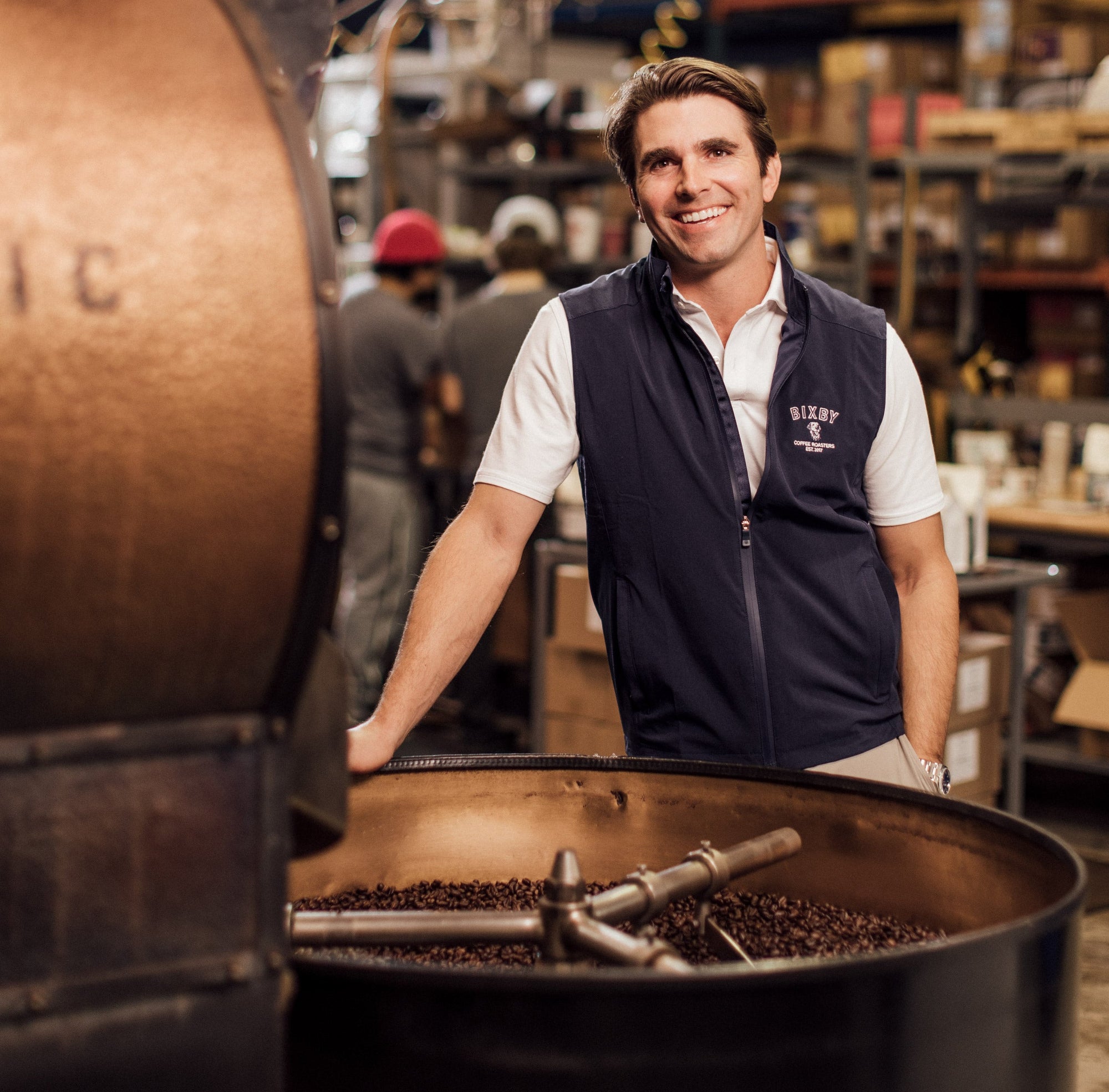 Smiling man in a navy vest over a white polo standing beside a large coffee roaster full of beans in a roastery.