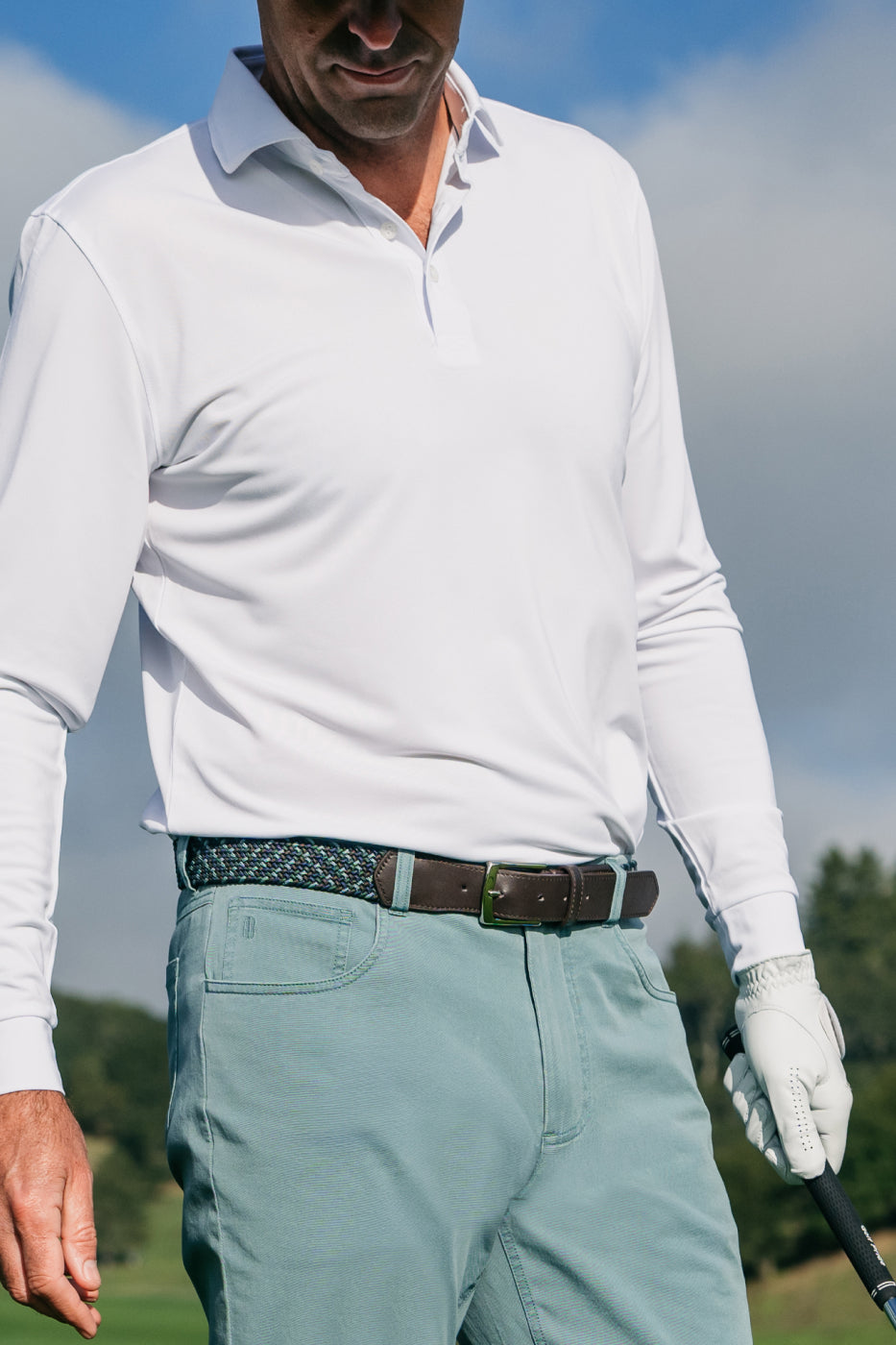 Golfer in a white long-sleeve polo, woven belt, sage green pants and a white golf glove holding a club grip.