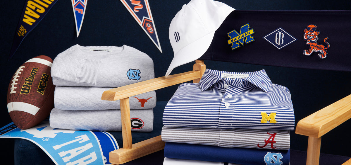 Folded sweaters and striped polos with embroidered college logos beside a white cap, football, pennants and a director's chair.
