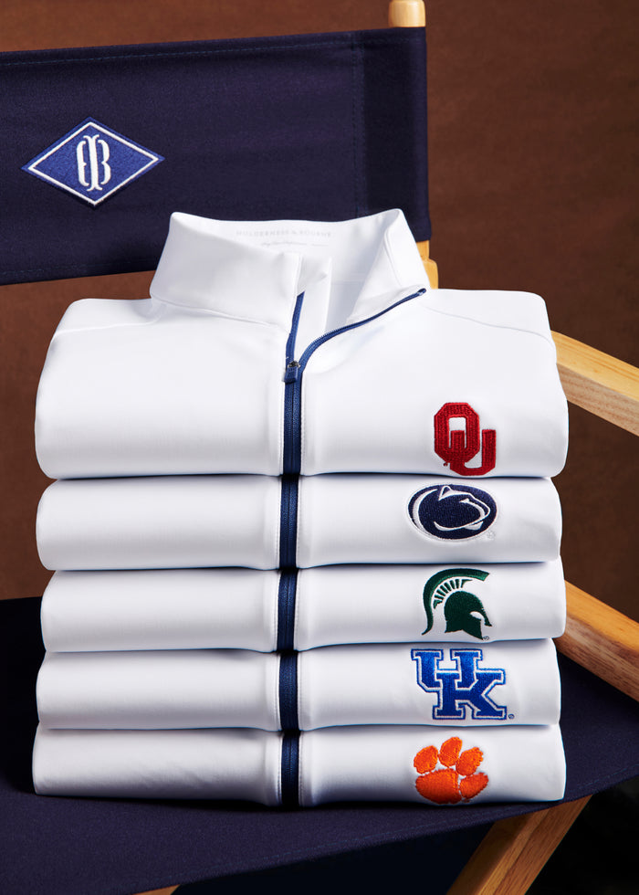 Stack of white quarter-zips with navy zippers embroidered with Oklahoma, Penn State, Michigan State, Kentucky and Clemson logos on a director's chair.