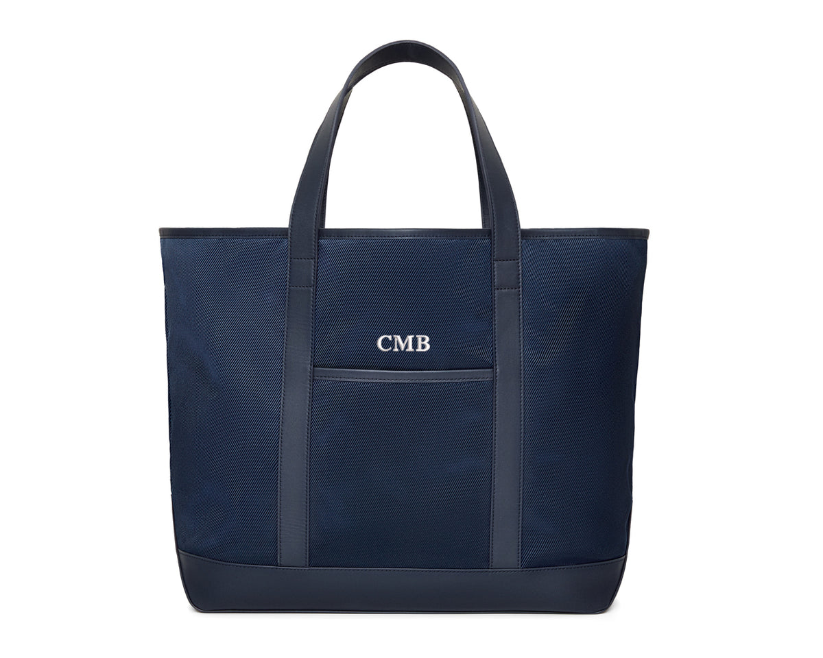 The Treadwell Tote: Navy Ballistic with white CMB monogram, features leather-trimmed handles, front pocket, and an open-top design for versatile use.