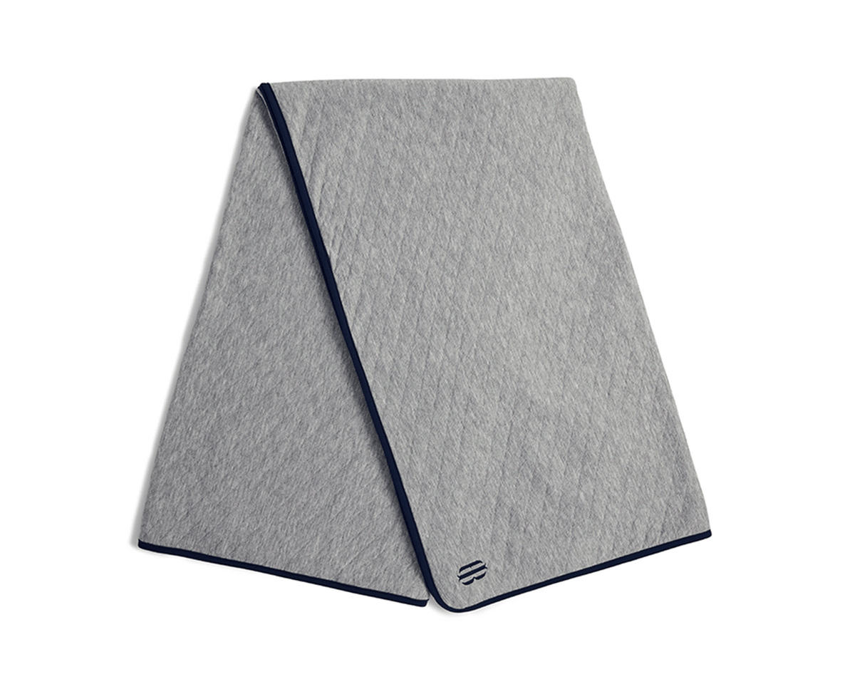The Van Pelt Blanket: Heathered Gray, a light-grey quilted blanket with diamond stitching, dark-navy trim, and a minimalist logo embroidered near one corner.