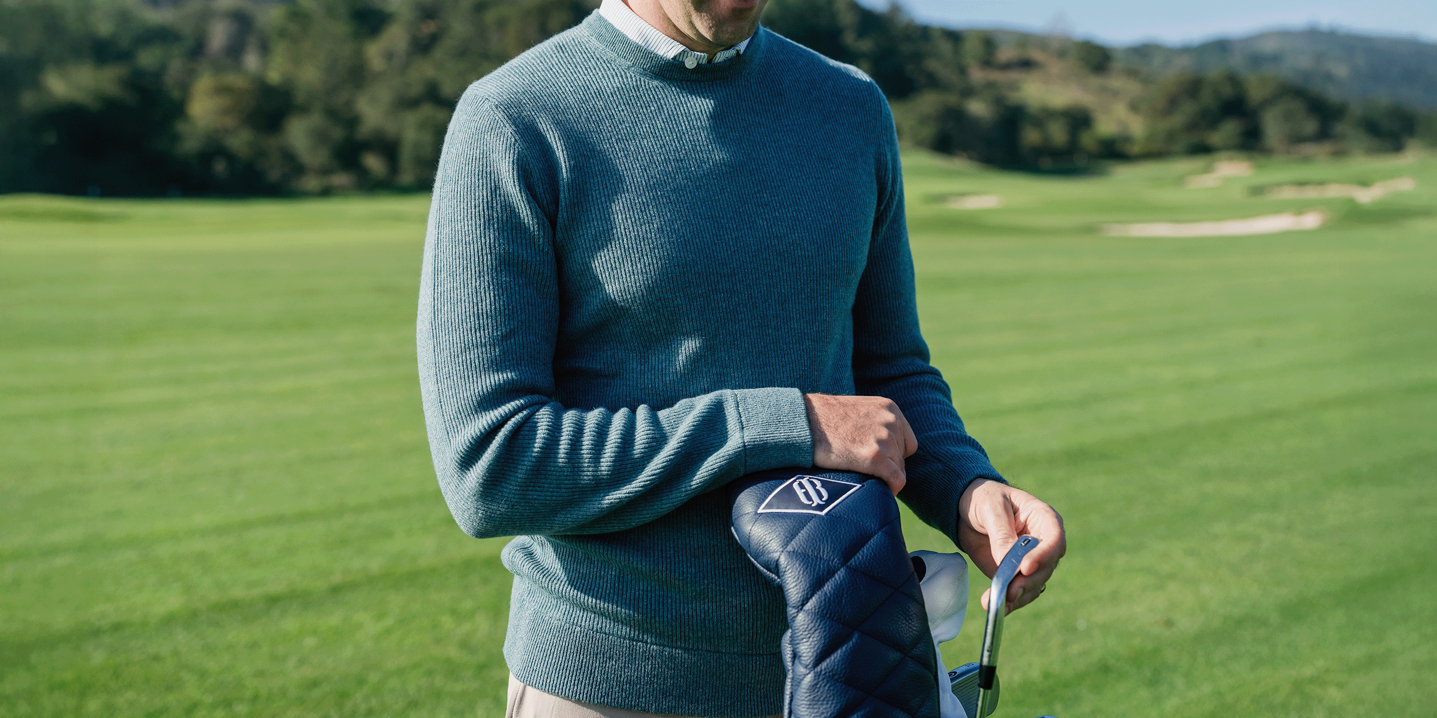 Golfer in a blue-gray crewneck sweater over a collared shirt holding a navy quilted headcover on a fairway.