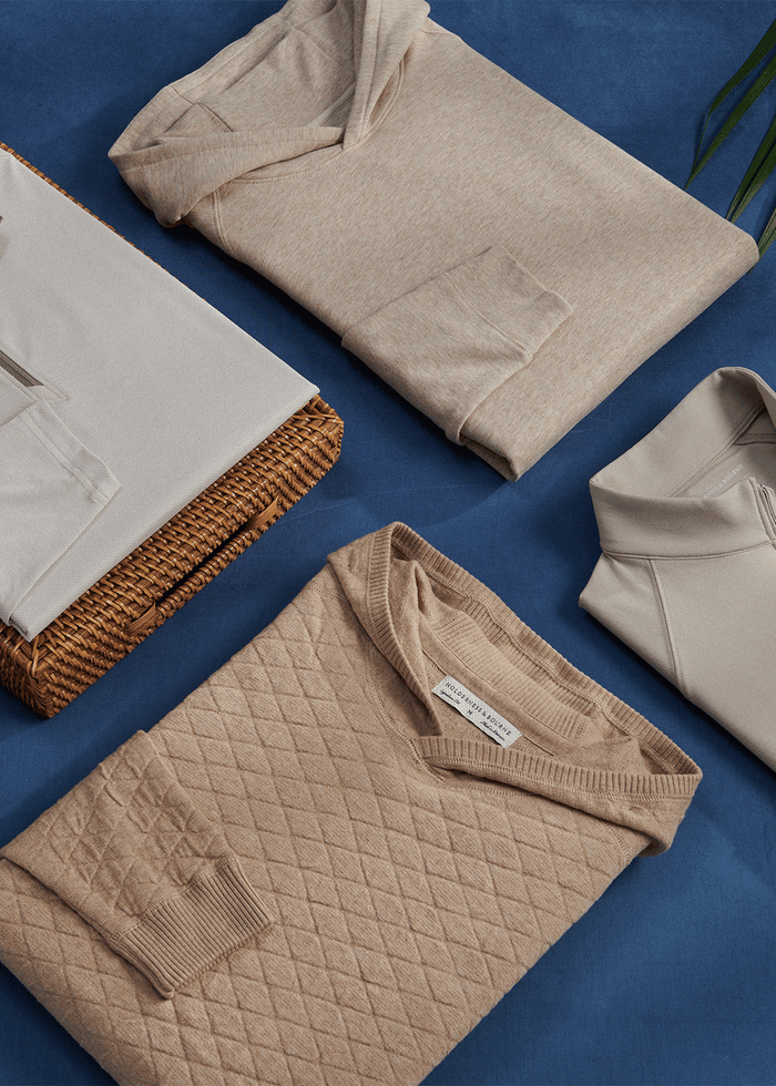 Flat lay of a quilted tan sweater, a hooded pullover and cream quarter-zips folded on blue cloth and a rattan tray.