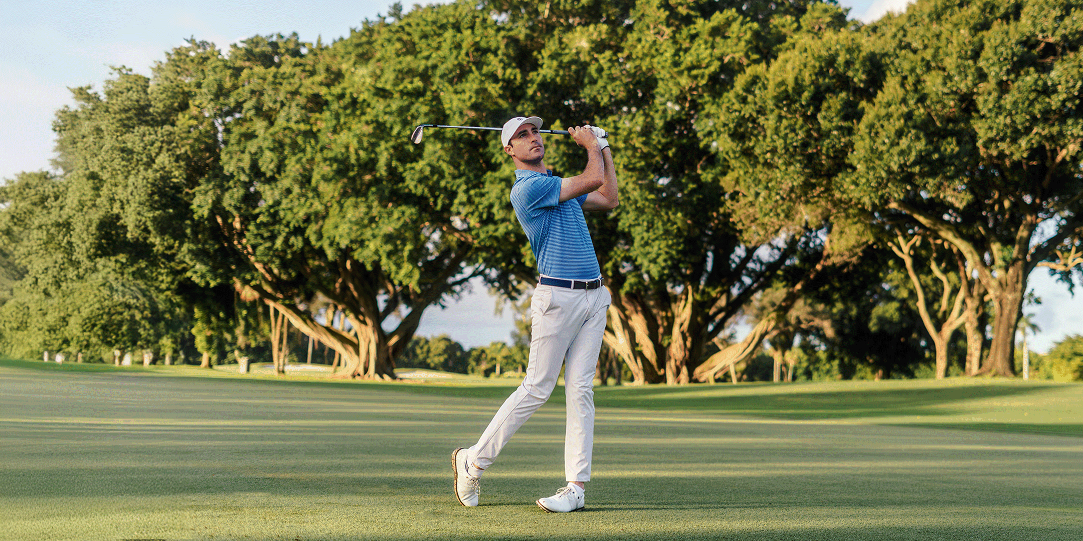 Golfer in blue shirt and white pants swinging a golf club on a green course with trees in the background