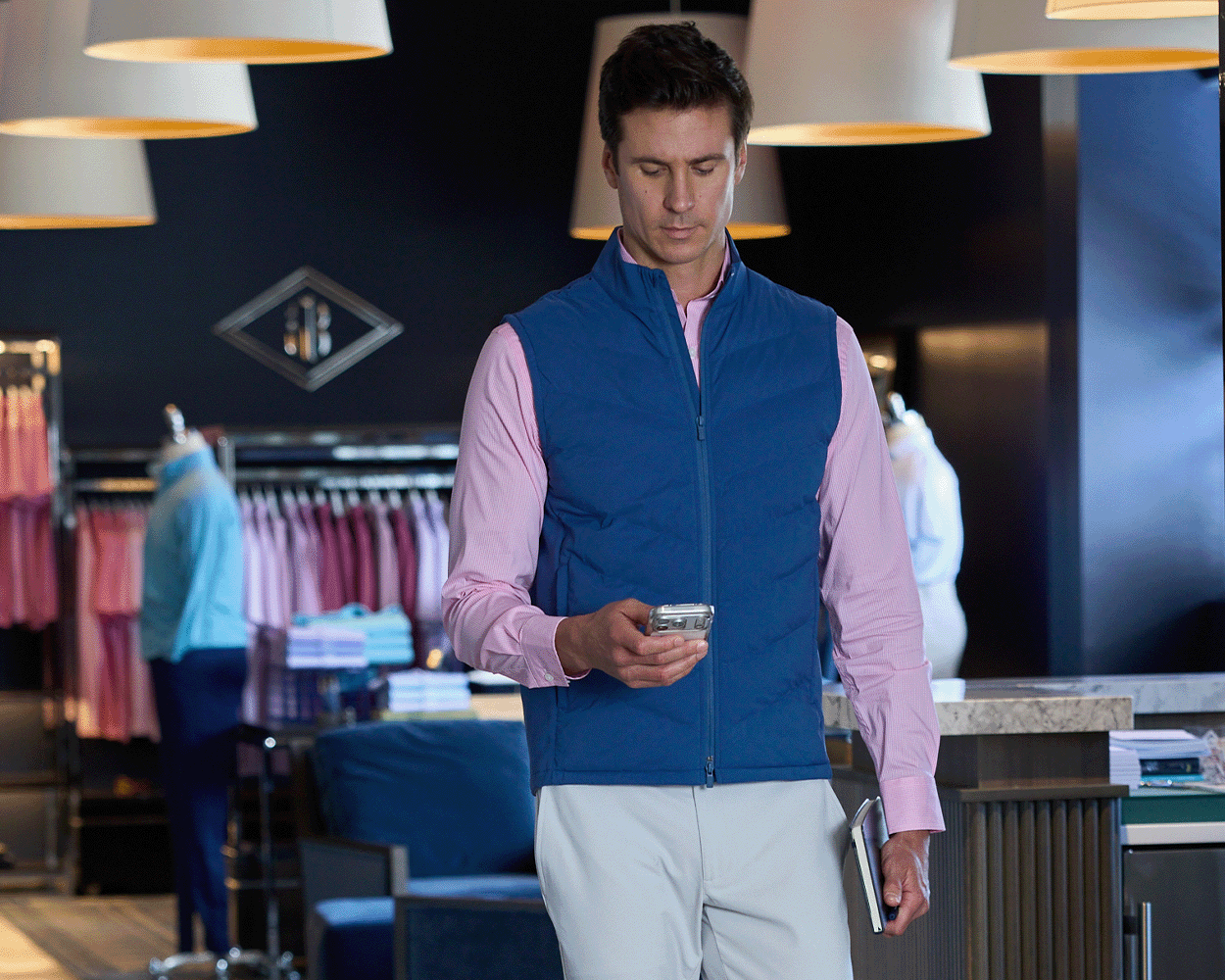 Man wearing The Benton Vest: Andover, a quilted vest with Thermore® insulation, stands in a boutique holding a smartphone and tablet, surrounded by clothing displays.