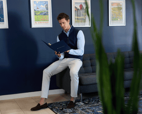 Man in The Cullinane Shirt: Heathered Vista Blue, seated on a sofa, leafing through a booklet, with vintage golf posters in the background.