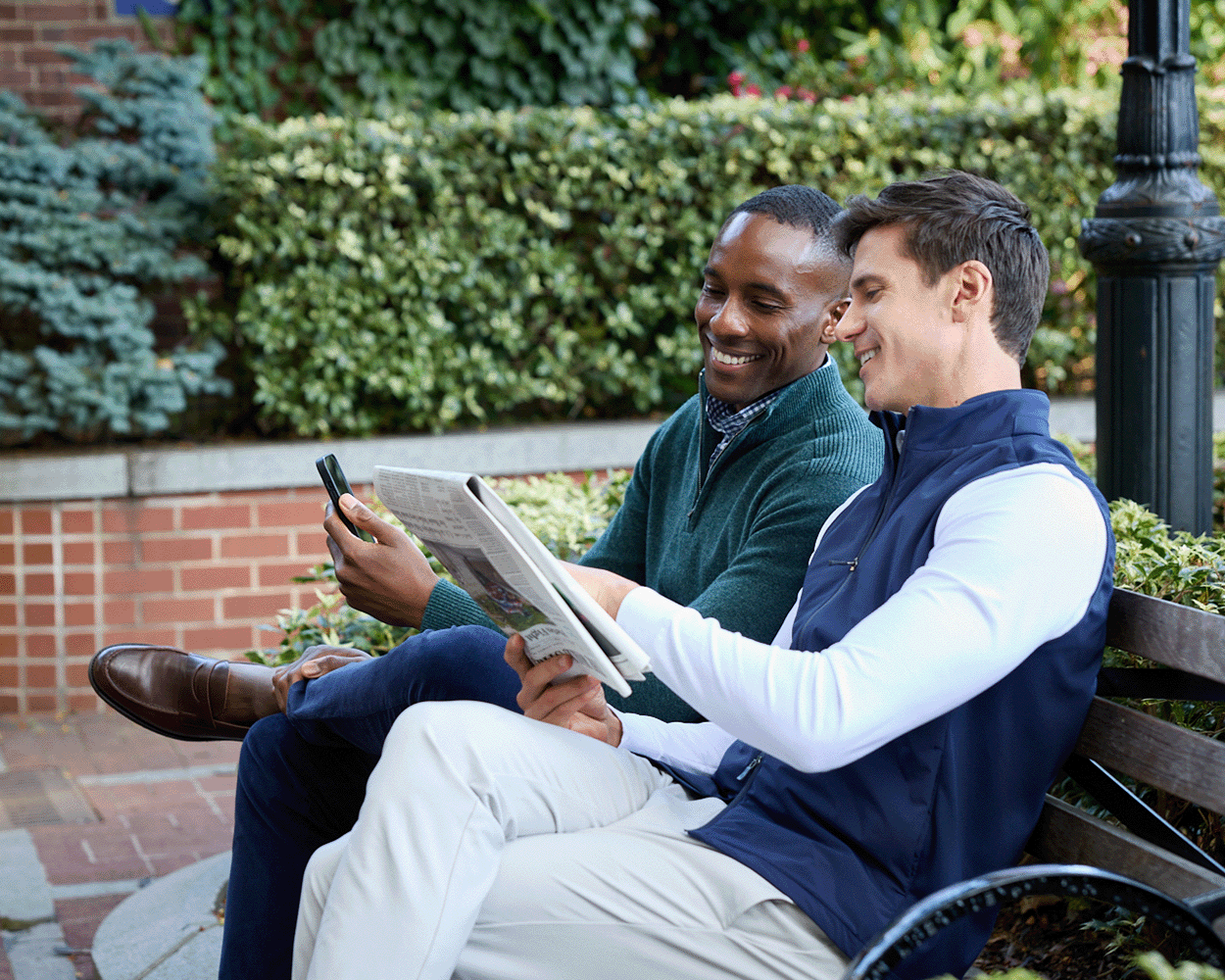 Two men sitting on a bench, smiling at a smartphone. One wears The Swenson Vest: Navy, showcasing its sleek design and functional warmth.