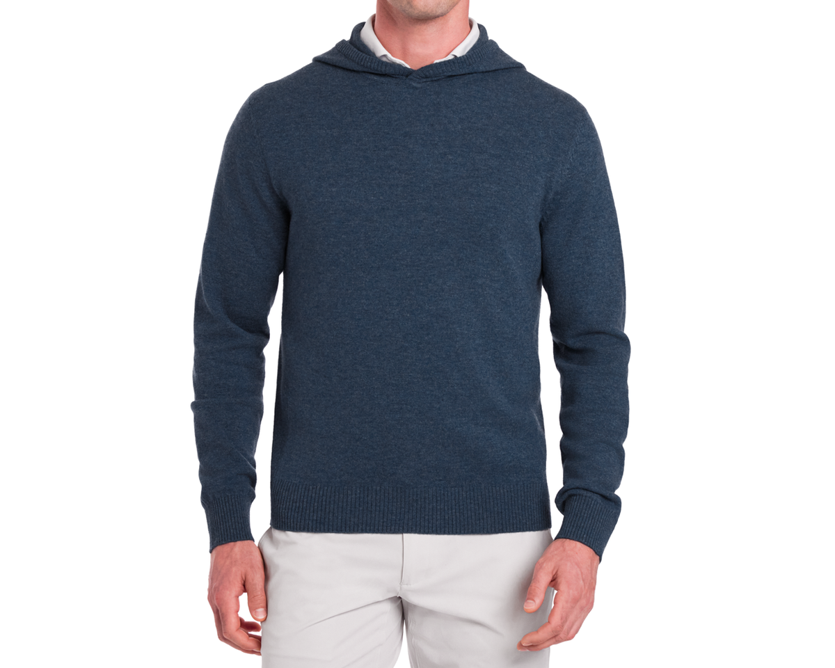 The Logan Hoodie Sweater: Heathered Andover, a navy-blue knit pullover with a hood, worn over a white shirt and paired with beige trousers.