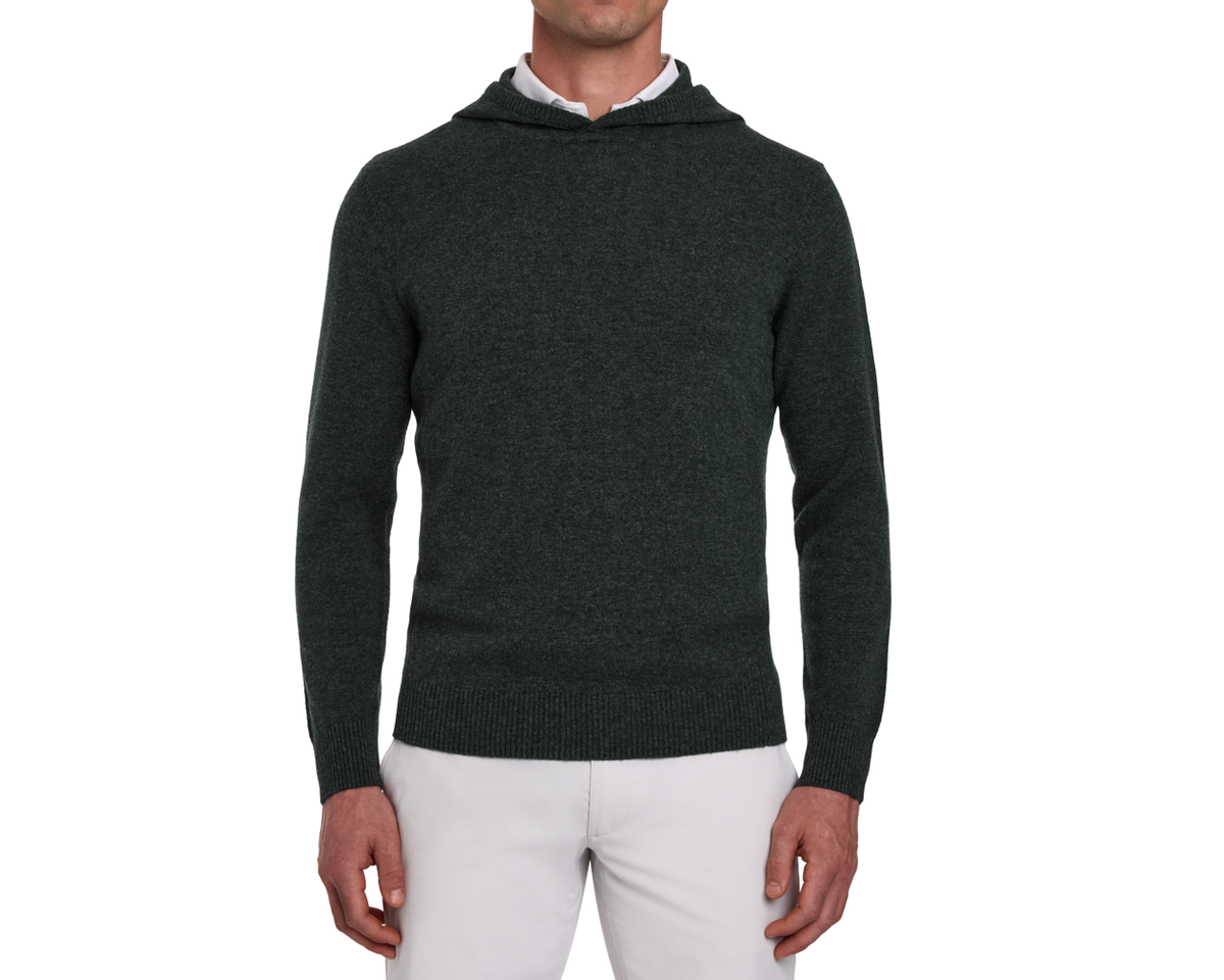 Man wearing The Logan Hoodie Sweater: Heathered Ivy, a knit hoodie over a white shirt, paired with light-gray pants, showcasing its minimalist design and ribbed details.