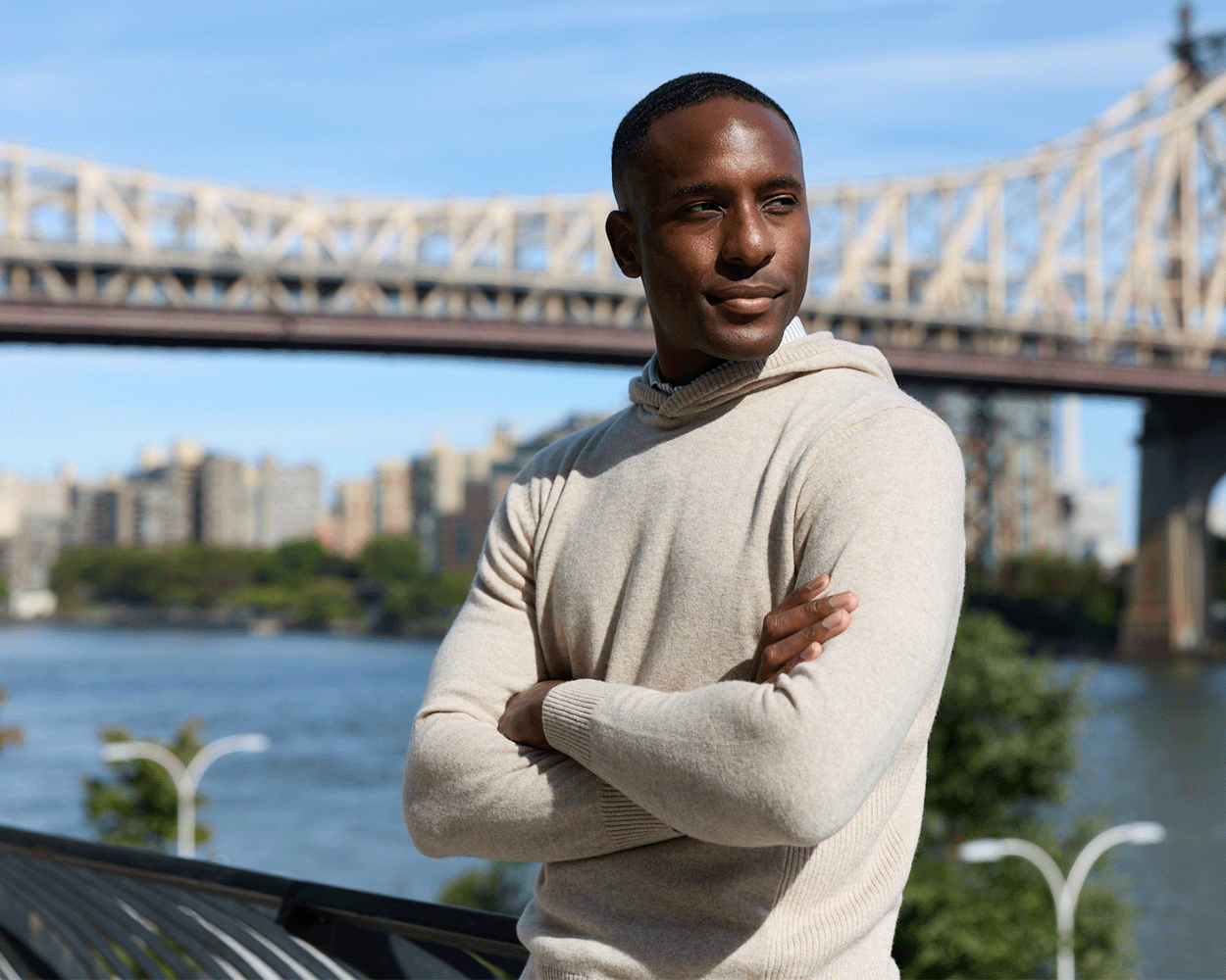 Young man wearing The Logan Hoodie Sweater in Heathered Dune stands on a waterfront promenade, with a bridge and cityscape in the background.