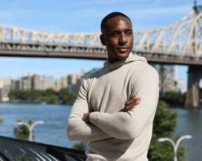 Young man wearing The Logan Hoodie Sweater in Heathered Dune stands on a waterfront promenade, with a bridge and cityscape in the background.