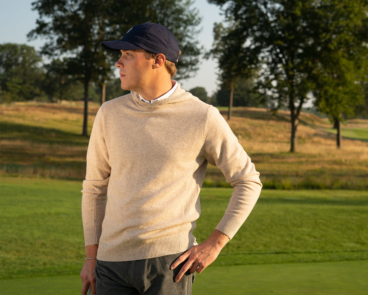 The Logan Hoodie Sweater: Heathered Dune, worn by a man on a golf course, showcases its luxurious blend and minimalist design in a relaxed, elegant setting.
