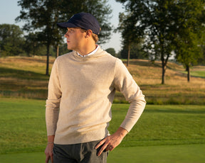 The Logan Hoodie Sweater: Heathered Dune, worn by a man on a golf course, showcases its luxurious blend and minimalist design in a relaxed, elegant setting.