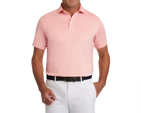 The Perkins Shirt: Nectarine & White, a light salmon-pink polo with short sleeves, tucked into white pants, featuring a dark woven belt and structured collar.