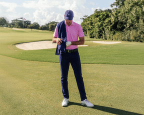 Man on golf course wearing The Maxwell Shirt: Belmont & White, adjusting golf glove. Features navy trousers, white golf shoes, navy cap, and performance pique fabric.