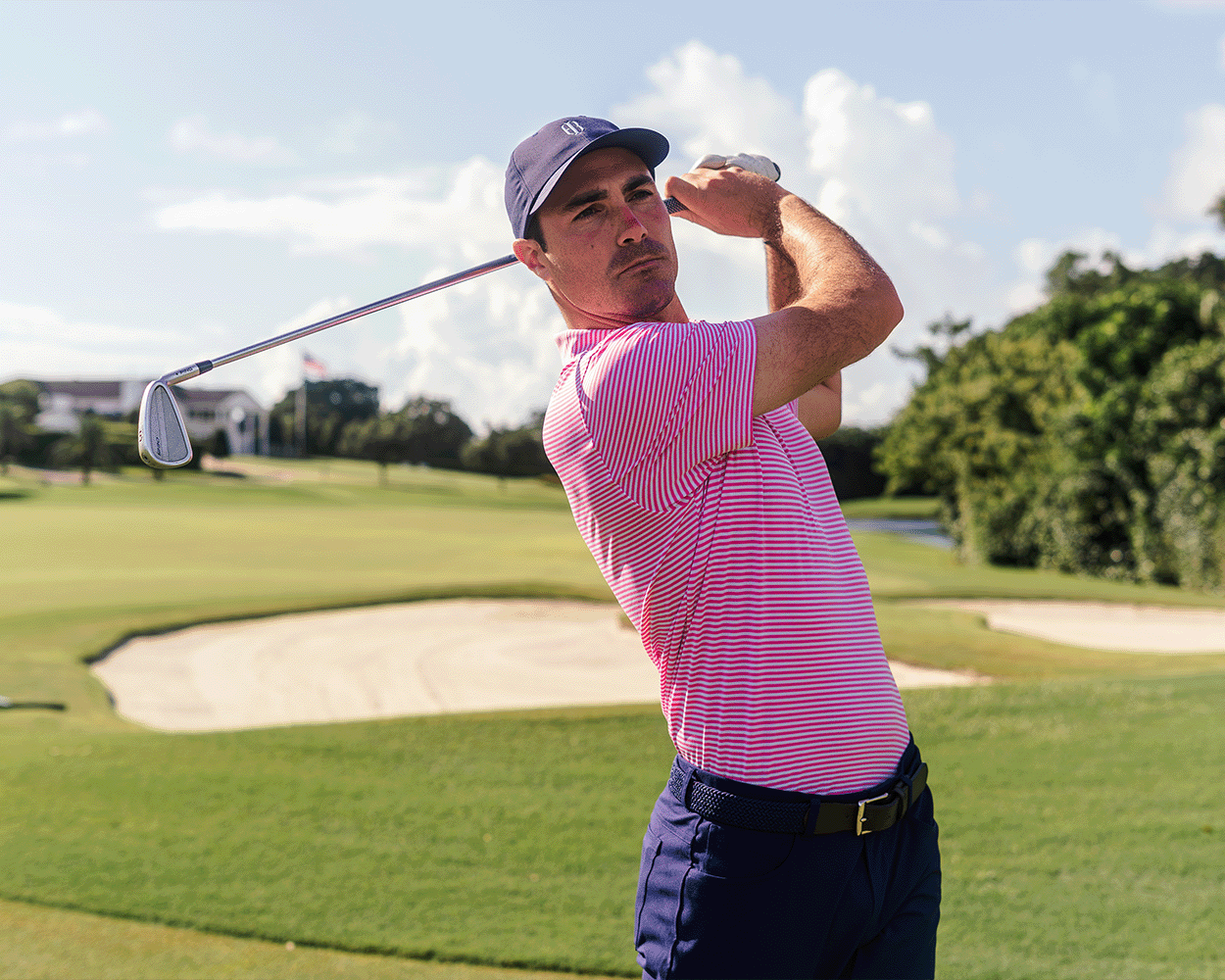 Man on golf course wearing The Maxwell Shirt: Belmont & White, showcasing a follow-through swing with an iron club, emphasizing the polo's comfort and performance features.