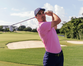Man on golf course wearing The Maxwell Shirt: Belmont & White, showcasing a follow-through swing with an iron club, emphasizing the polo's comfort and performance features.